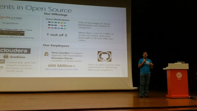 peaker on a stage presenting an open source slide deck projected on a large screen, with the person’s face blurred