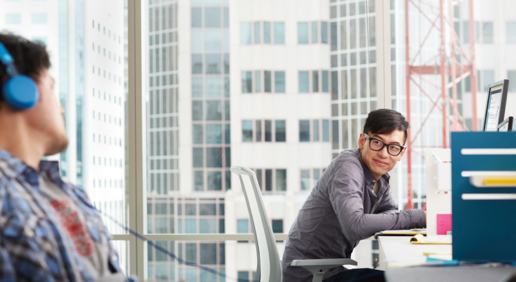 Two coworkers sit at desks in a bright office with floor‑to‑ceiling windows