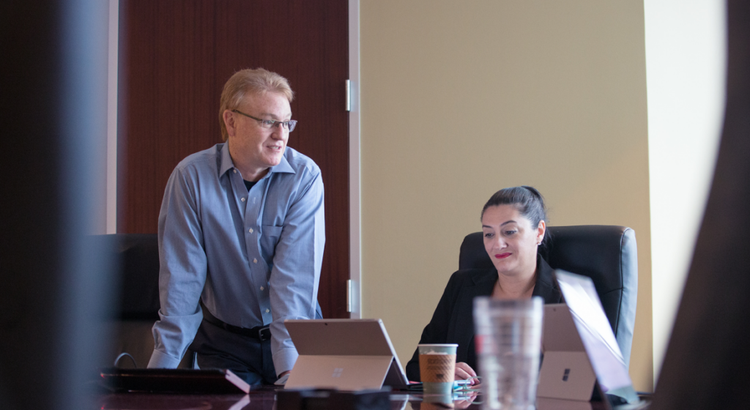 A man in a light blue shirt leans over a conference table next to a seated woman with a laptop