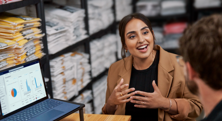 Woman in a tan blazer enthusiastically explains data shown on a laptop screen