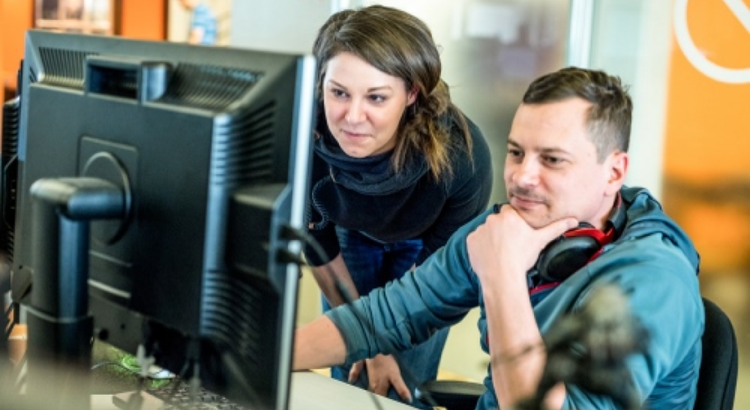 Two coworkers closely review something on a large desktop monitor