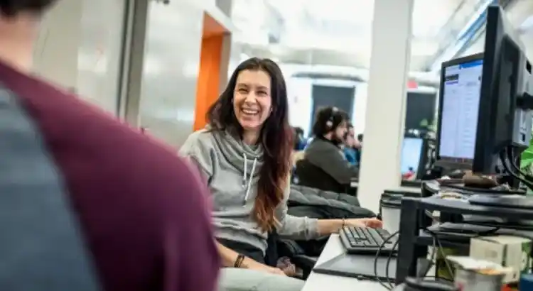 A woman in a gray hoodie sits at a desk with a computer monitor,