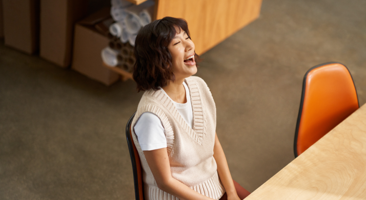 Woman sitting at a wooden table in a workspace leans back laughing