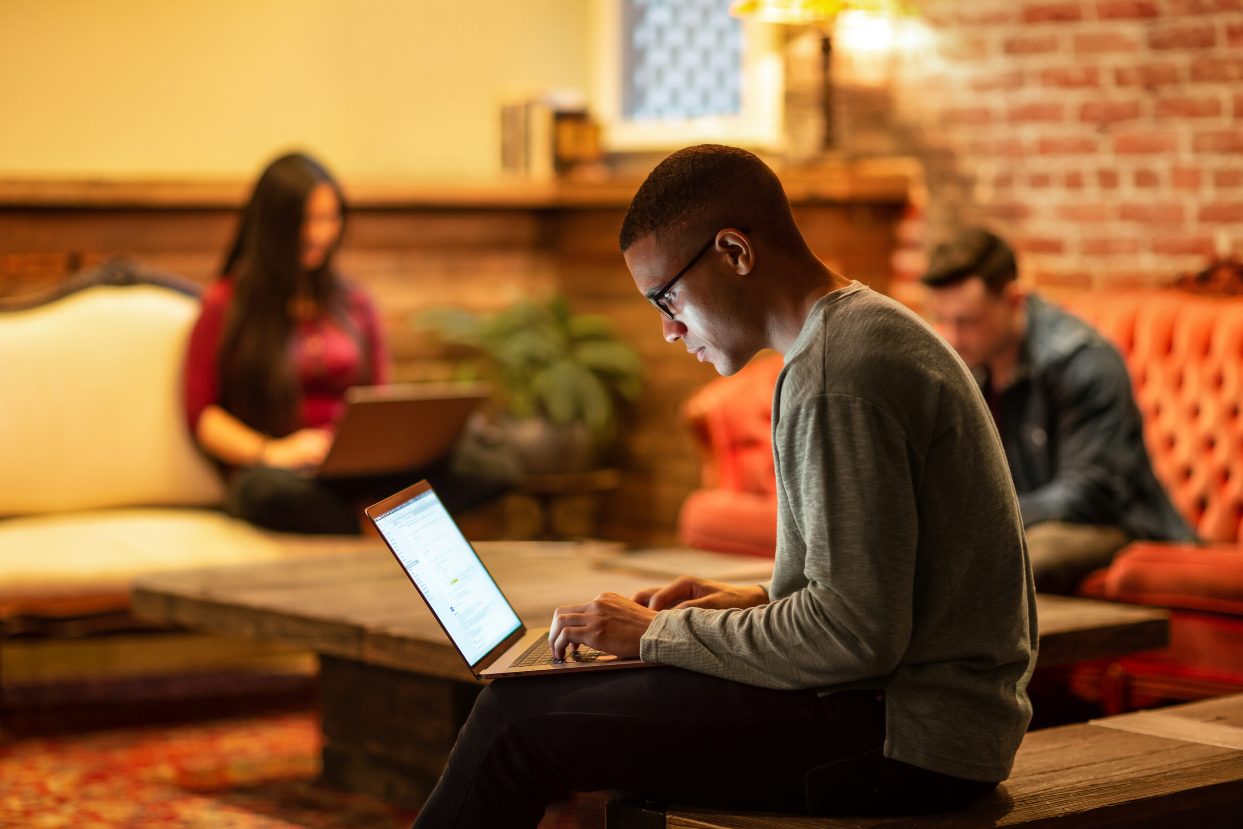 Person wearing glasses works on a laptop in a café-style workspace, with two other people using laptops in the background