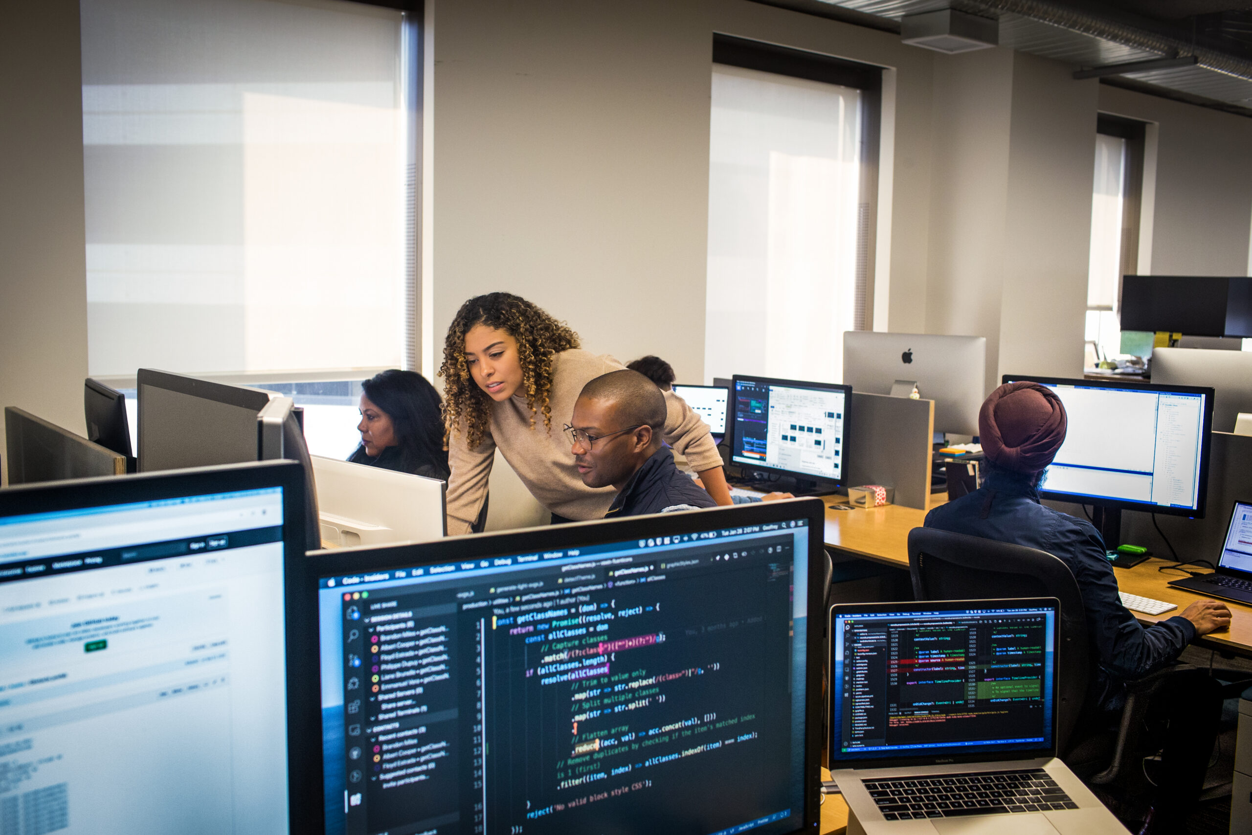 Software developers collaborate at computer workstations in an open office, with code displayed on multiple monitors and laptops